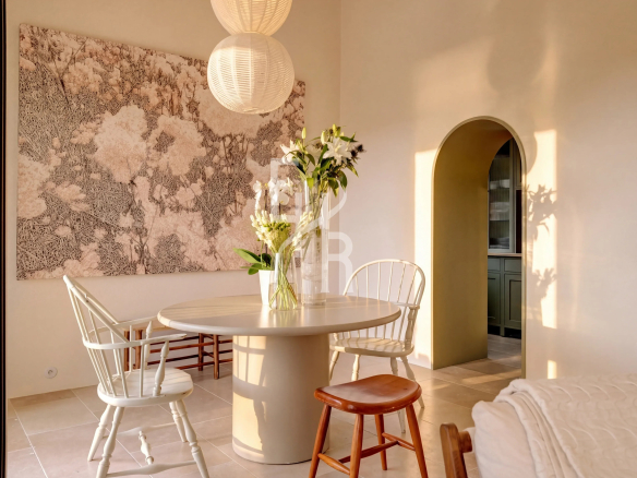 Interior of a bright dining room with a round table and chairs, decorated with flowers and a wall artwork.
