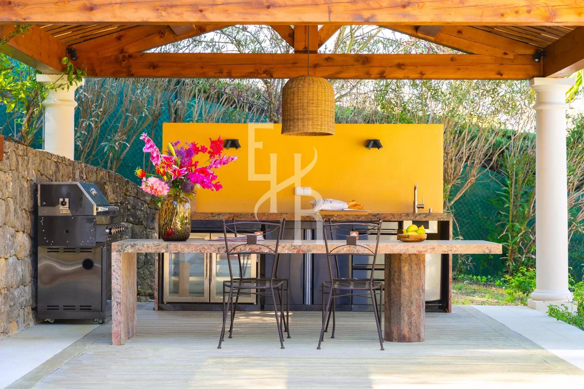 Cuisine extérieure avec table en pierre et chaises, fleurs en vase, sous un abri en bois à Mougins.