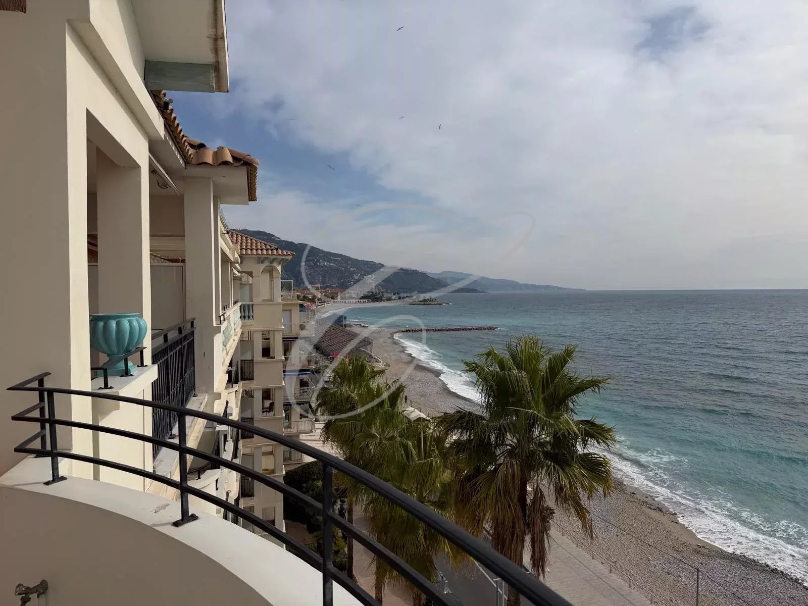 Balcony view of a Mediterranean-style hotel overlooking a pebbly beach and turquoise sea.