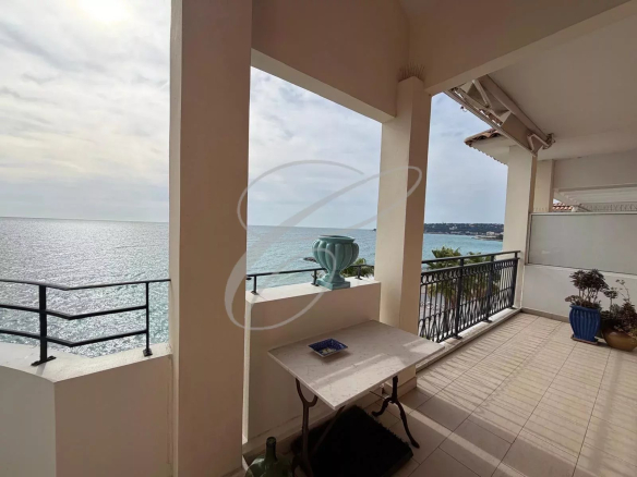 Balcony overlooking the sea with a rail, beige pillars, a stone table, and potted plants by the edge, calm ocean in the background