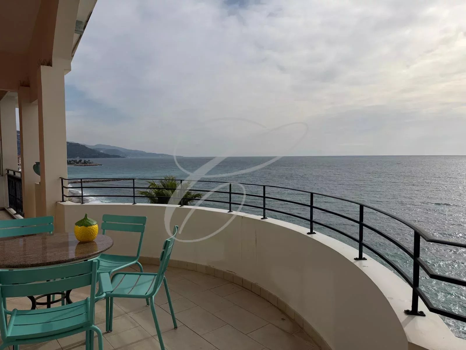 Ocean view from a curved balcony with turquoise chairs and a round table topped by a yellow decorative item, overlooking the sea.