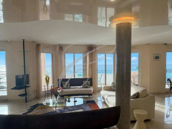Bright seaside living room with white sofas, glass coffee table, and floor-to-ceiling windows overlooking the ocean on a sunny day, with a balcony beyond the glass doors.