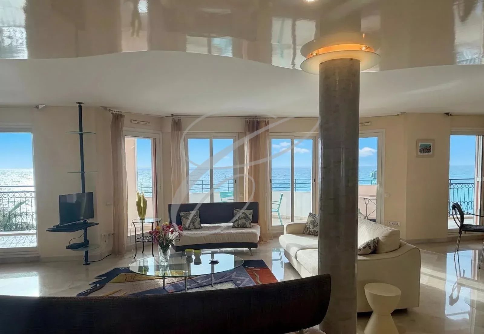 Bright seaside living room with white sofas, glass coffee table, and floor-to-ceiling windows overlooking the ocean on a sunny day, with a balcony beyond the glass doors.
