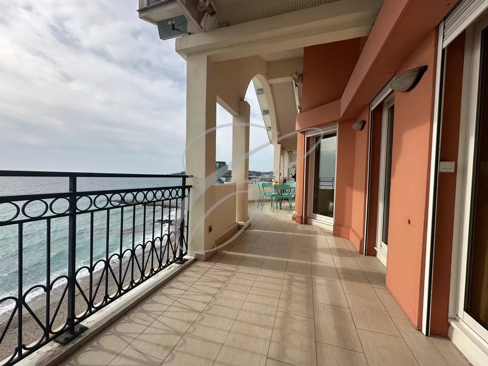Seaside balcony with a black wrought-iron railing, beige tiled floor, and orange building walls; turquoise chairs and table in the distance by the sea.