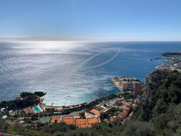 Coastal cliffside town with orange tennis courts and buildings along the shore under a bright blue sky.
