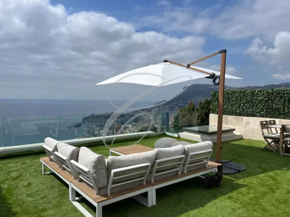 Outdoor rooftop lounge with white cushioned sectional sofa, a large white umbrella, and an ocean view.