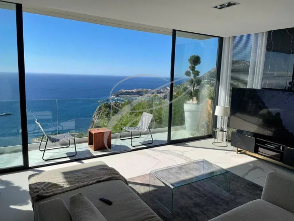 Modern living room with floor-to-ceiling glass walls overlooking the blue ocean and cliffside views; two patio chairs on a glass balcony.