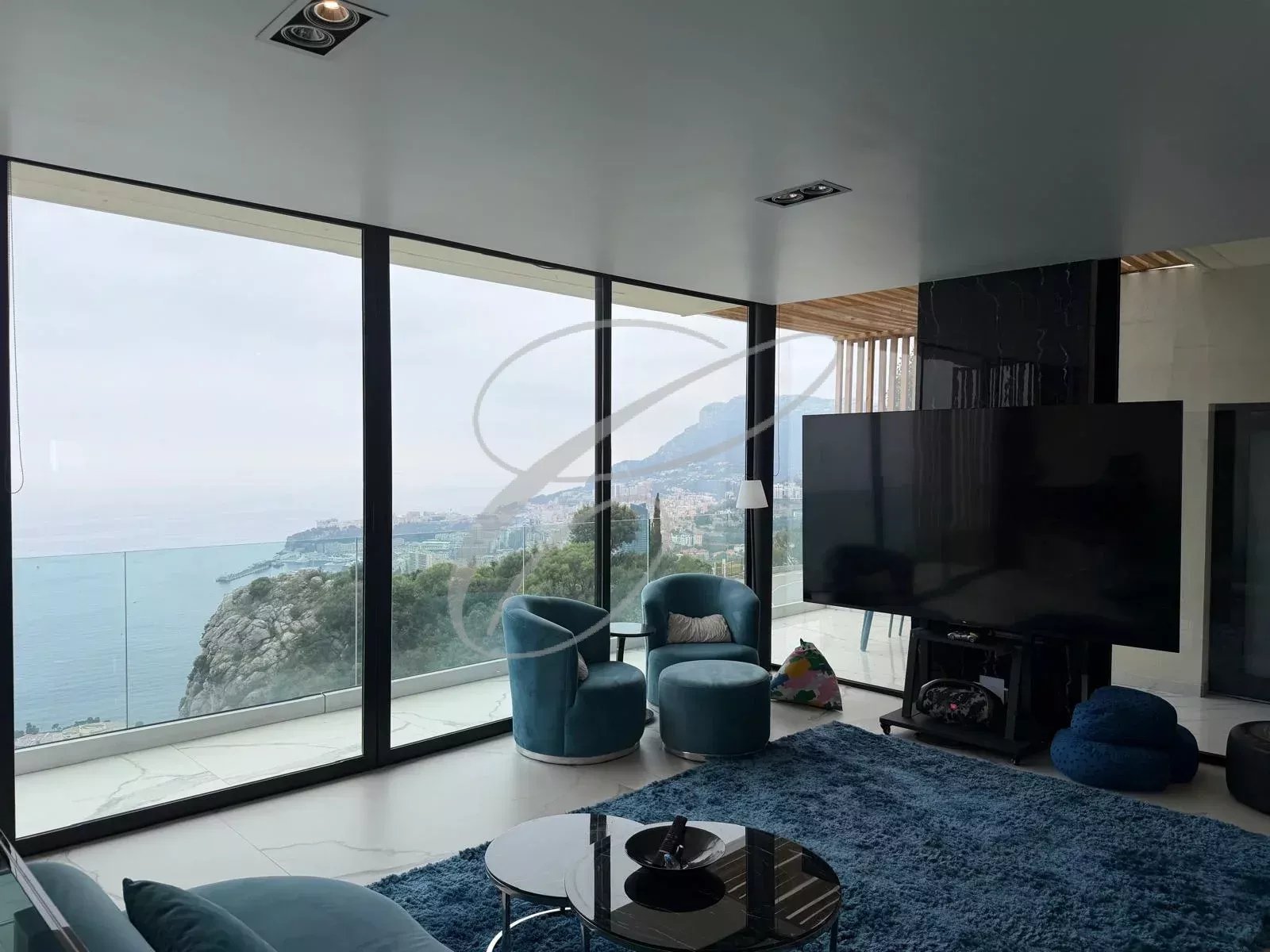 Modern living room with floor-to-ceiling windows showing a coastal view and hillside town outside.