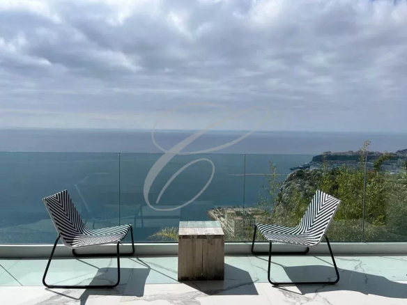 Two striped lounge chairs face a glass railing with a small wooden table between them, overlooking the sea and rocky coastline under a cloudy sky.