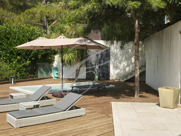 Terrasse d'une villa moderne avec jacuzzi, chaises longues et parasol, entourée de verdure.