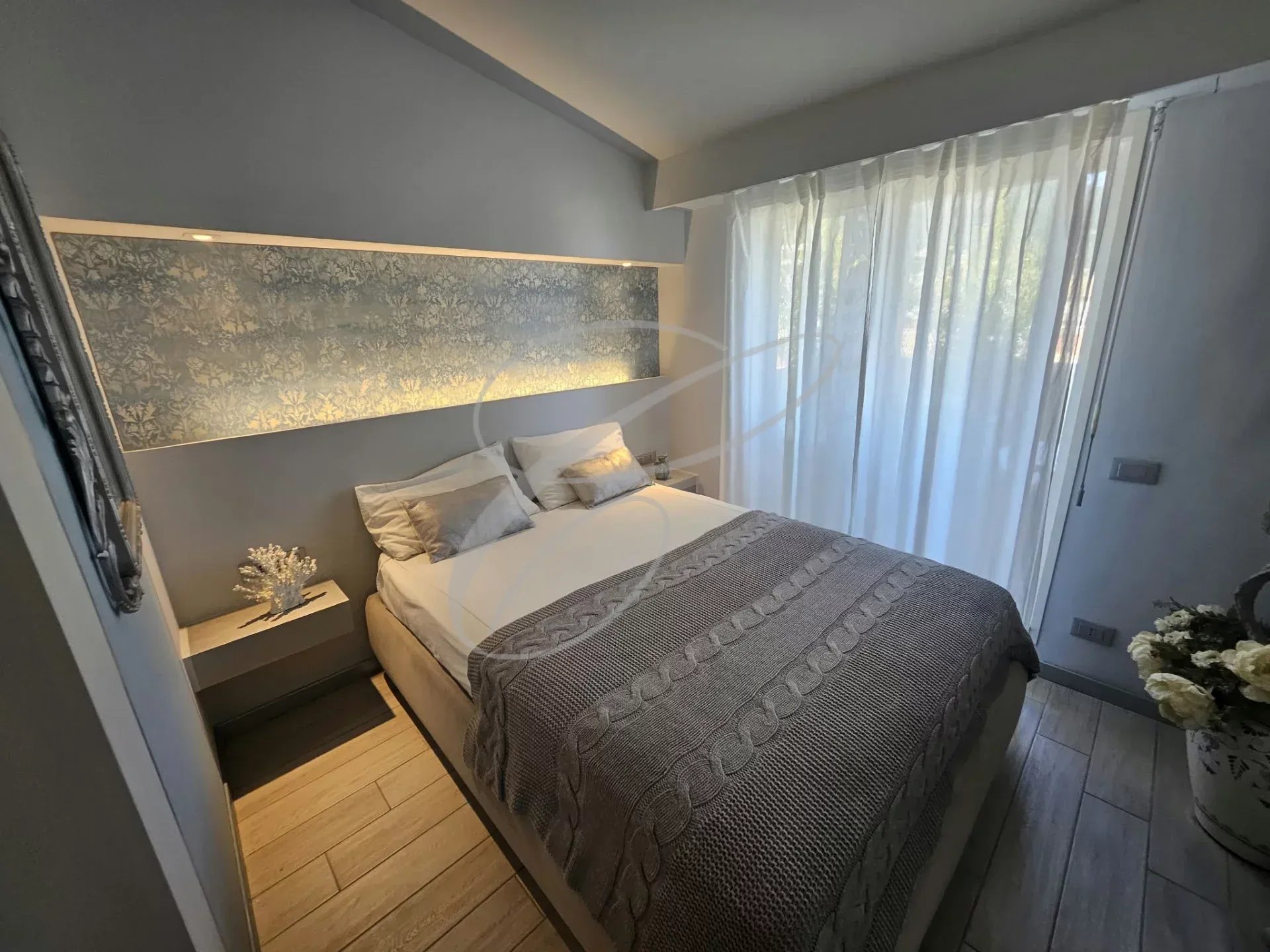 Chambre d'un appartement rénové avec lit double et fenêtre laissant entrer la lumière.