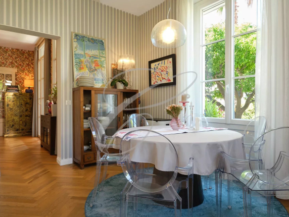 Salle à manger d'une villa avec table ronde et chaises transparentes, fenêtres donnant sur un jardin.