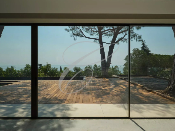 Vue depuis l'intérieur d'une villa contemporaine avec terrasse en bois et arbres, surplombant la mer.