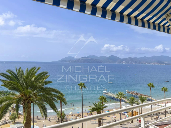 Vue panoramique sur la mer depuis un balcon avec des palmiers et des parasols rayés à Cannes.