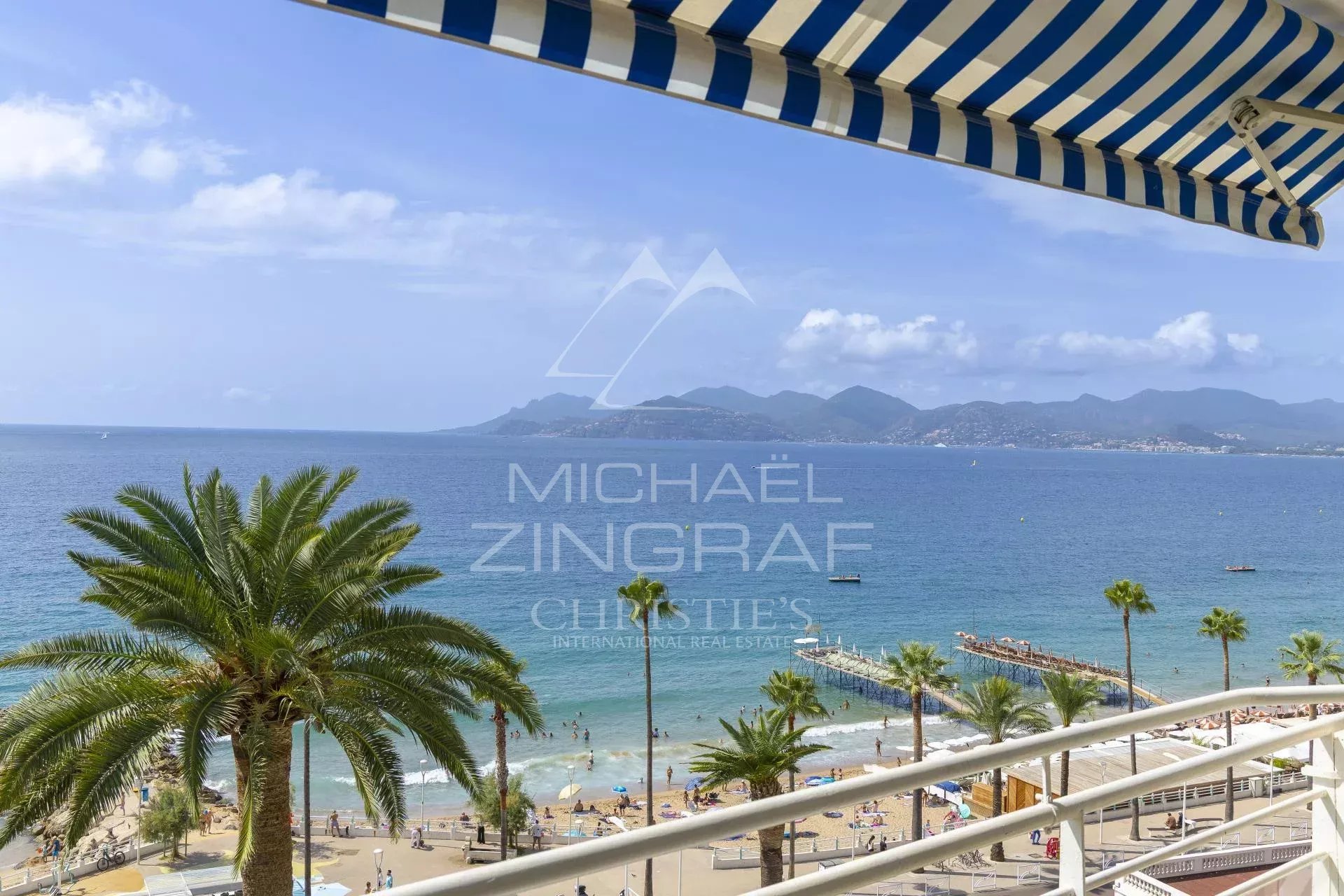 Vue panoramique sur la mer depuis un balcon avec des palmiers et des parasols rayés à Cannes.