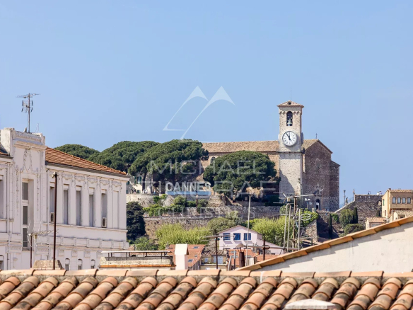 Vue sur une église avec horloge et collines à Cannes, entre des bâtiments en bordure de toit.