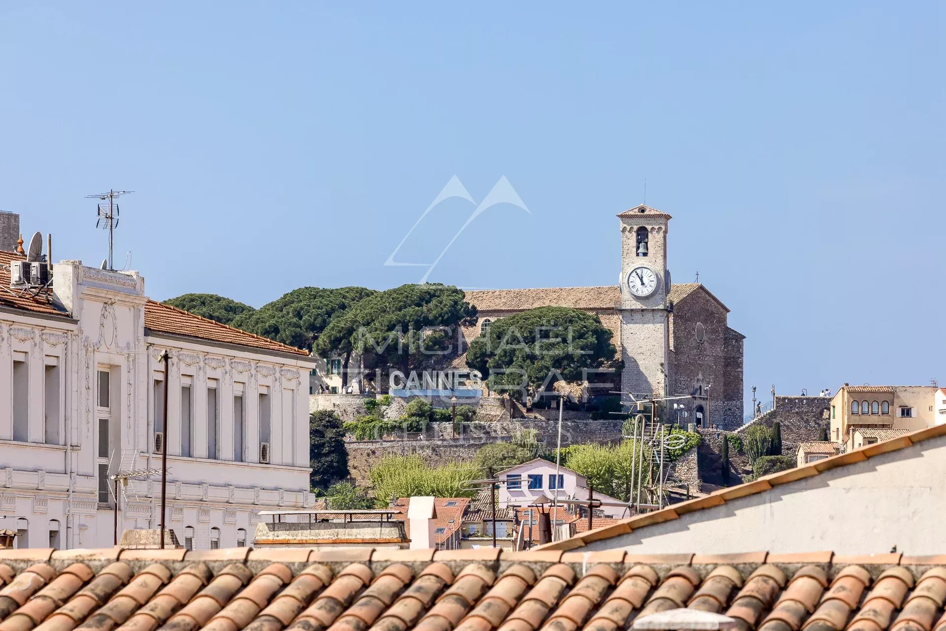 Vue sur l’église de Cannes Vue sur une église avec horloge et collines à Cannes, entre des bâtiments en bordure de toit.