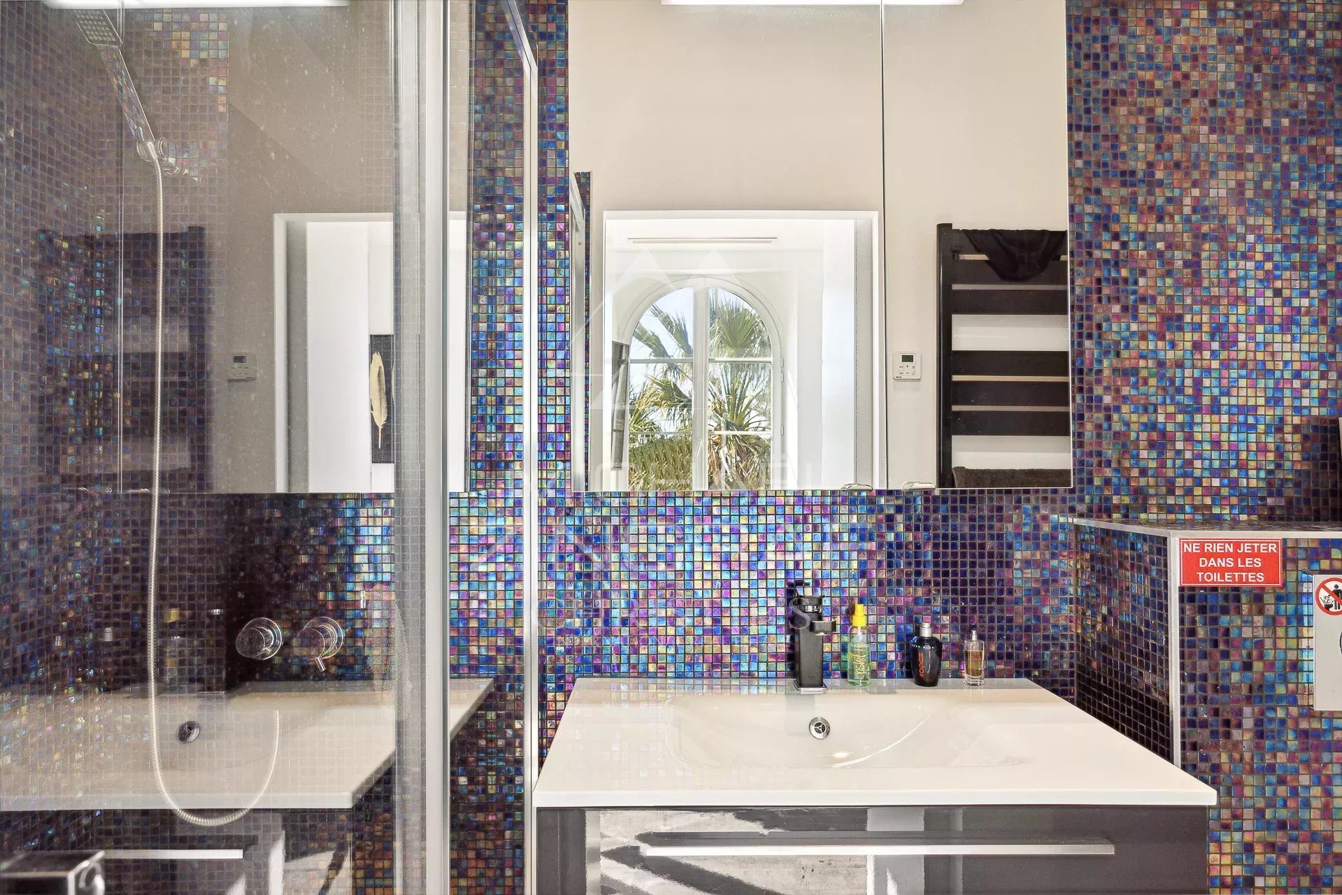 Salle de bain du penthouse à Cannes Salle de bain moderne avec carrelage coloré et miroir, vue sur une fenêtre avec palmier.