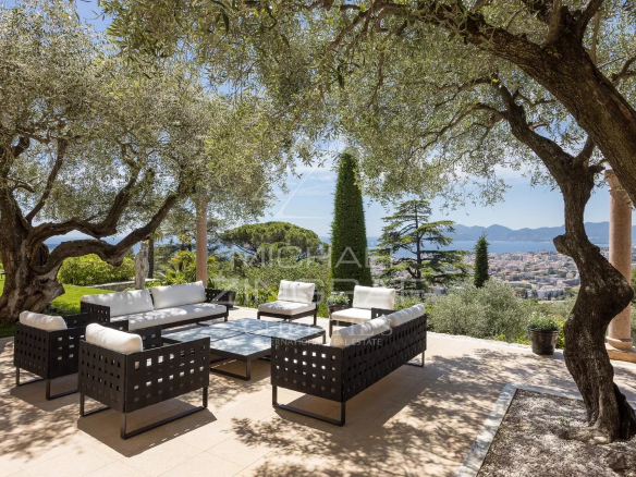 Terrasse avec mobilier moderne et vue sur la baie de Cannes, entourée d'arbres et de jardins paysagers.