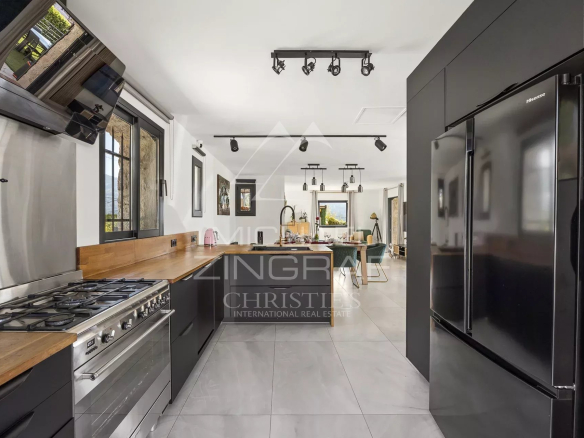 Open-plan kitchen with dark cabinetry, wooden countertops, stainless steel gas range and large fridge, leading to a dining/living area in the background.