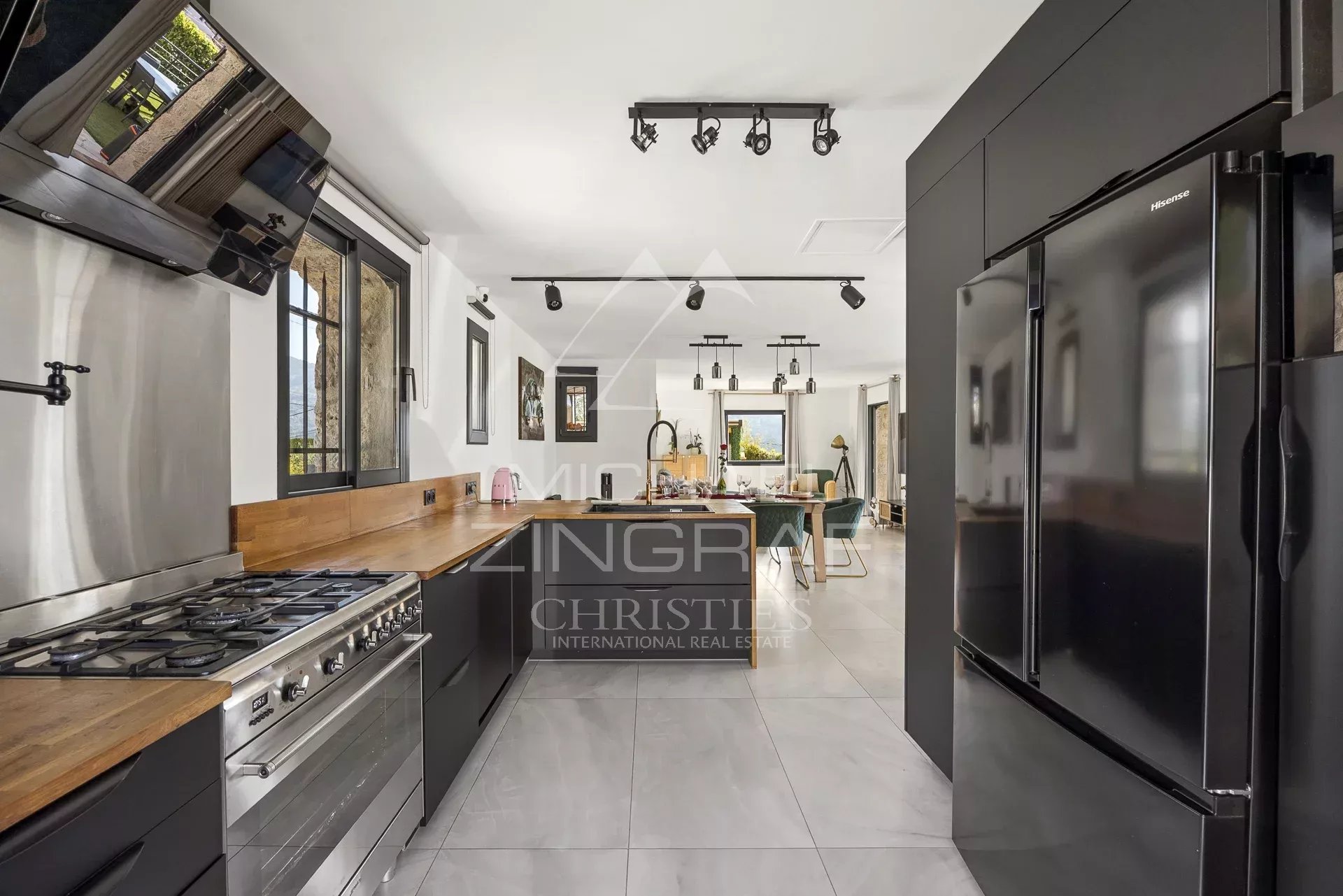 Open-plan kitchen with dark cabinetry, wooden countertops, stainless steel gas range and large fridge, leading to a dining/living area in the background.