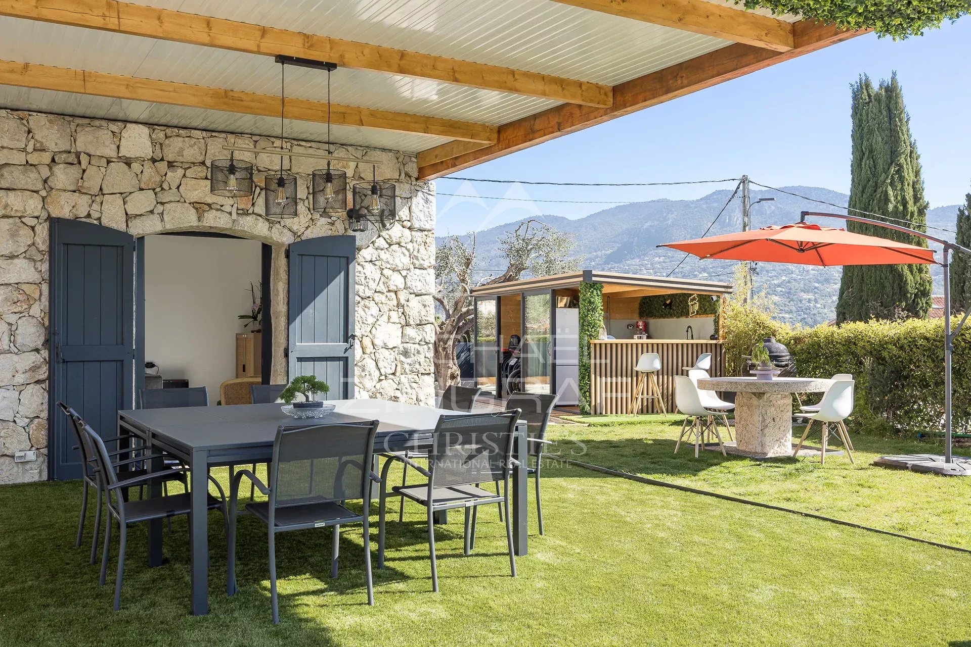 Stone wall house with blue doors opening to a covered patio and a large outdoor dining table on the lawn