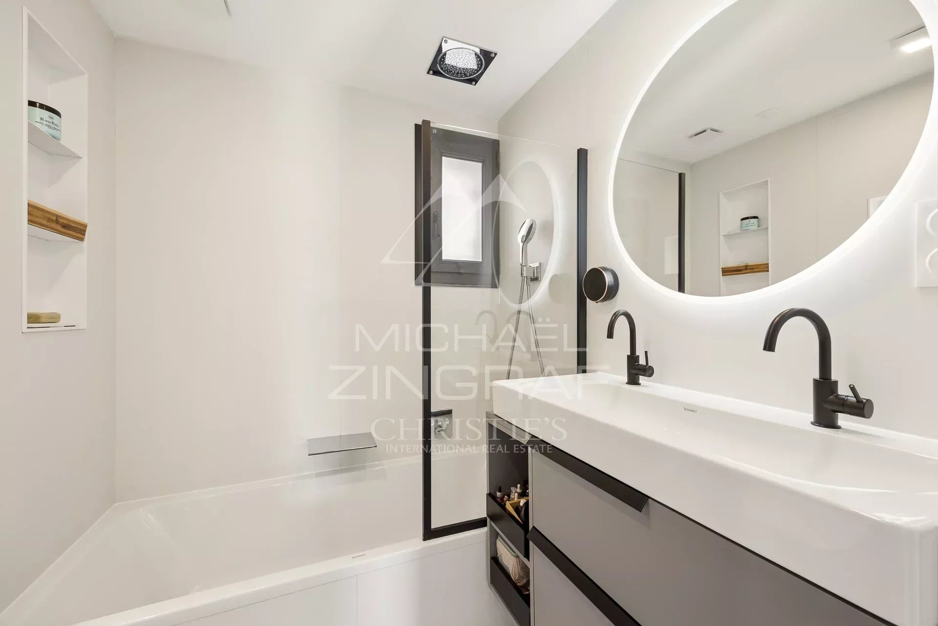 Modern white bathroom with a double sink, black faucets, and an illuminated round mirror above a grey vanity.