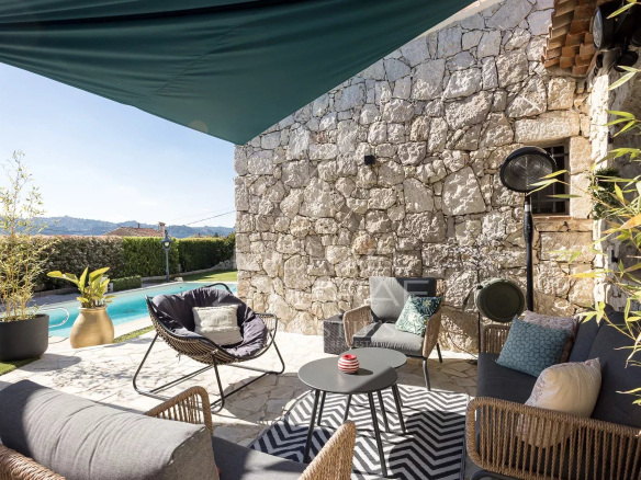 Outdoor lounge area beside a stone wall and pool, with woven sofas, cushions, and a round coffee table under a green canopy.