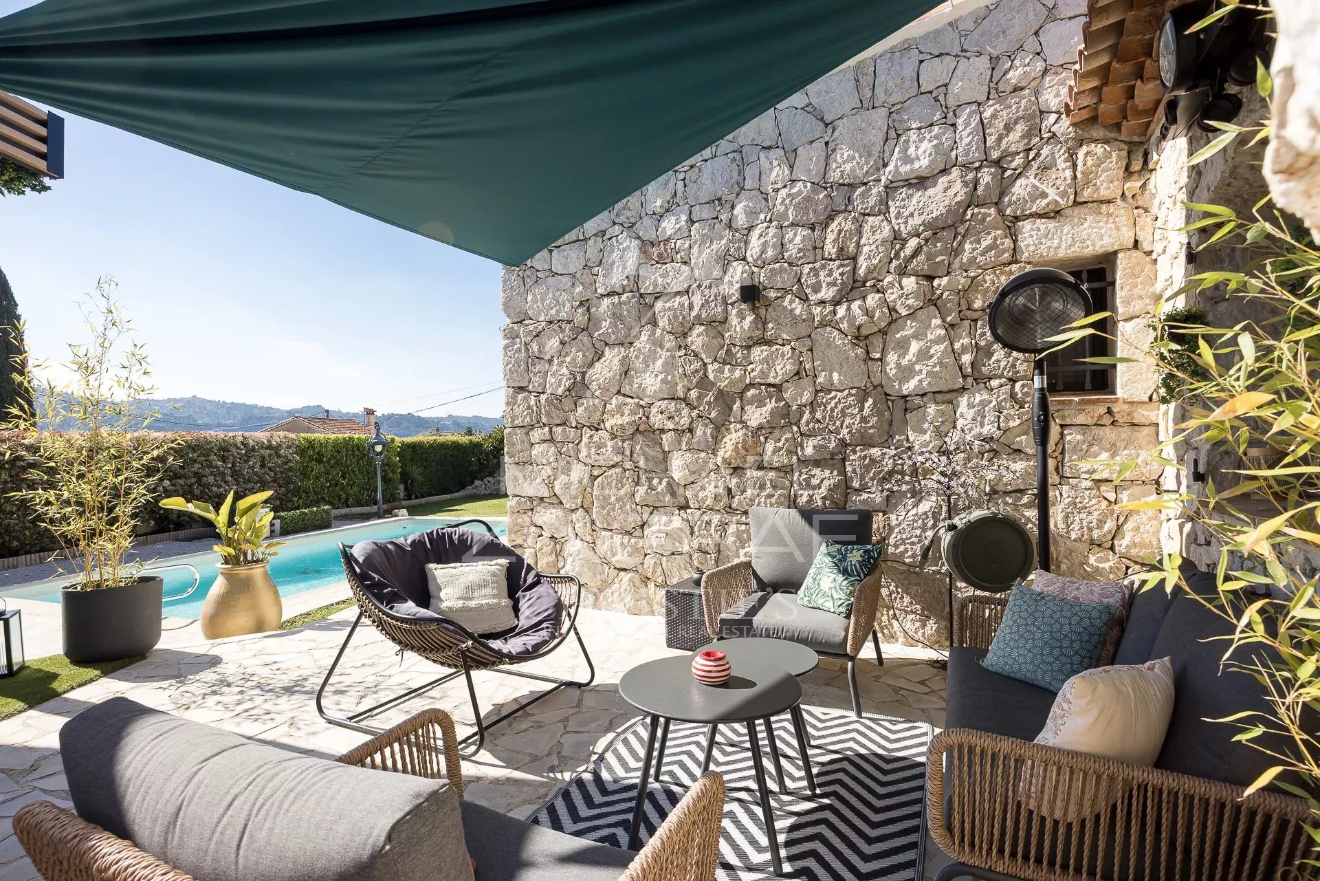 Outdoor lounge area beside a stone wall and pool, with woven sofas, cushions, and a round coffee table under a green canopy.