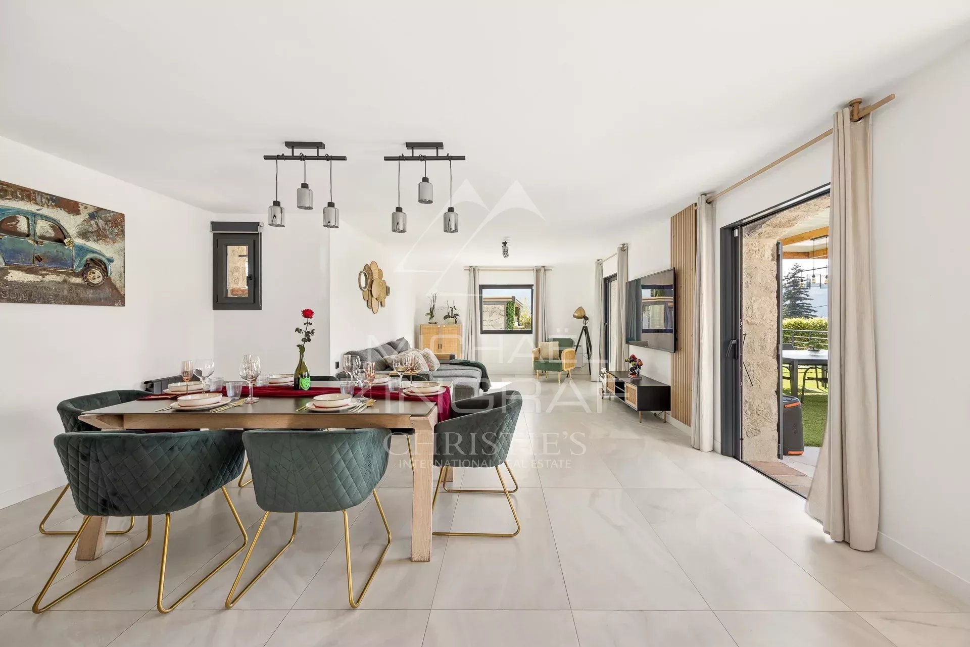 Open-plan dining and living area with a wooden table, six green quilted chairs, pendant lights, and sliding glass doors to a patio outside.