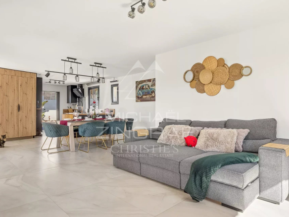 Open-plan living and dining area with a gray sectional, woven-basket wall art, and a wooden cabinet near the kitchen. A cat sits on the tiled floor left.