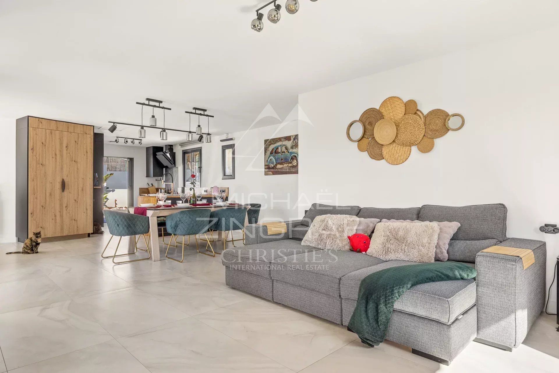Open-plan living and dining area with a gray sectional, woven-basket wall art, and a wooden cabinet near the kitchen. A cat sits on the tiled floor left.