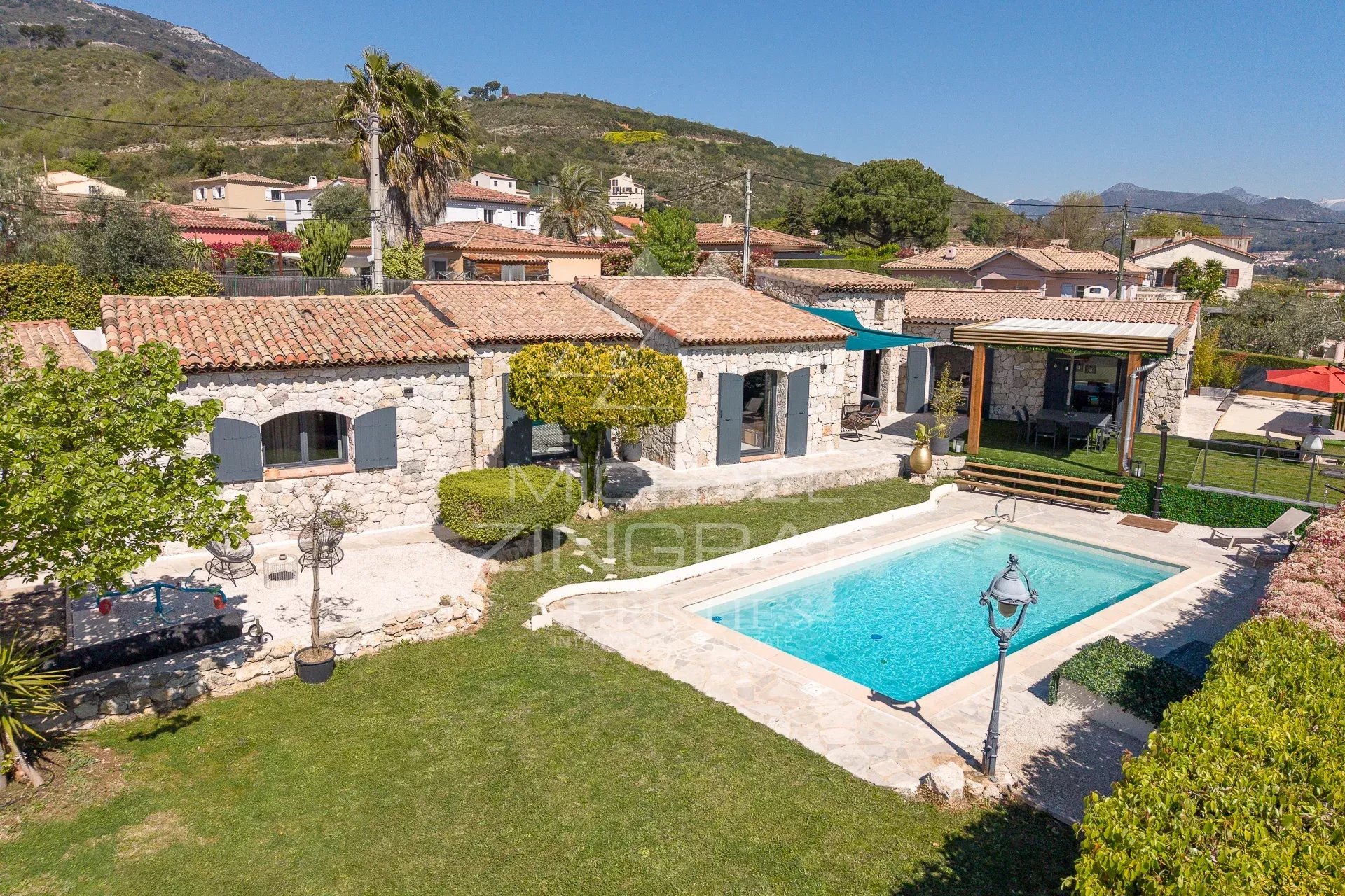 Stone villa with tiled roof and blue shutters, overlooking a garden and a rectangular pool area.