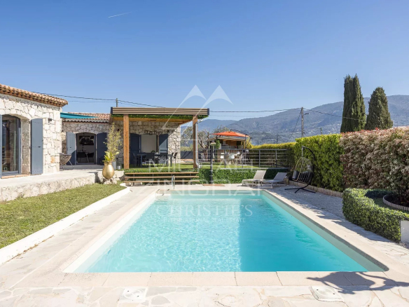 Stone house with blue shutters and a rectangular pool in a sunny yard with mountain backdrop