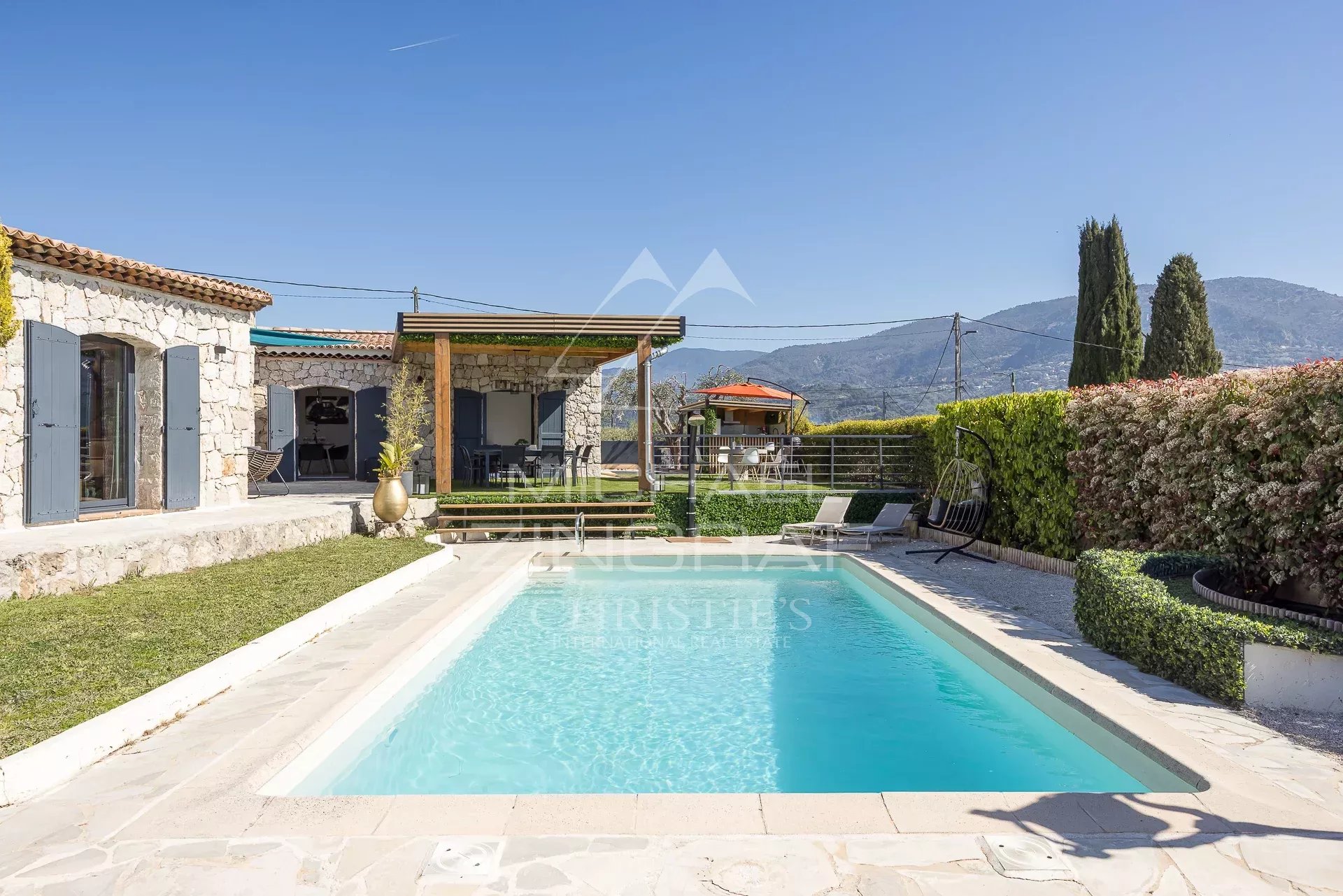 Stone house with blue shutters and a rectangular pool in a sunny yard with mountain backdrop
