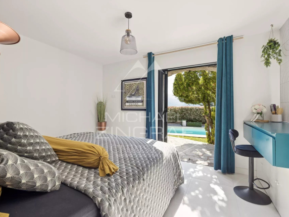 Bright bedroom with a patterned duvet, mustard pillows, and a blue desk by the window opening to a pool view outside.