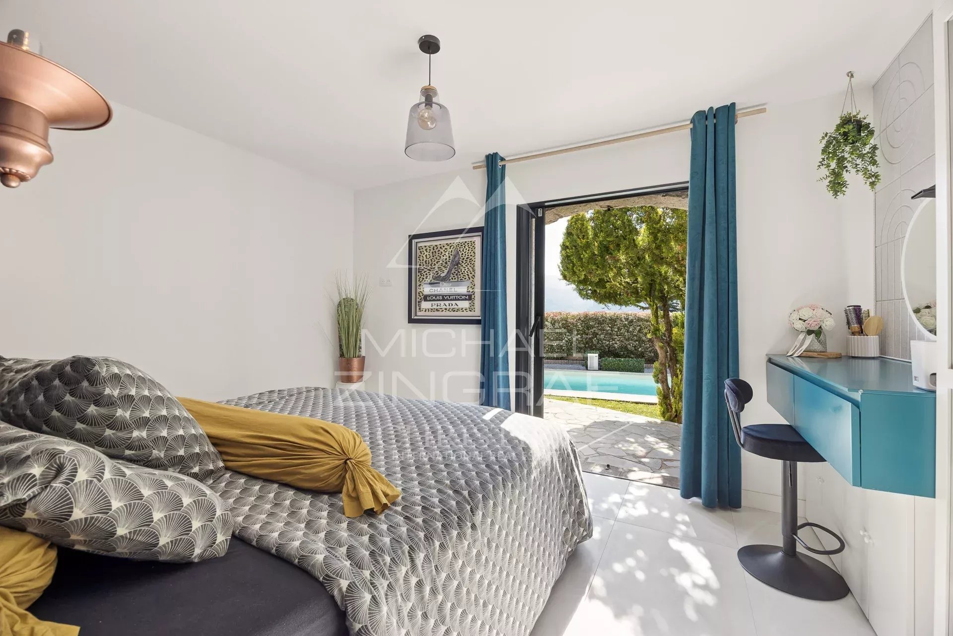 Bright bedroom with a patterned duvet, mustard pillows, and a blue desk by the window opening to a pool view outside.