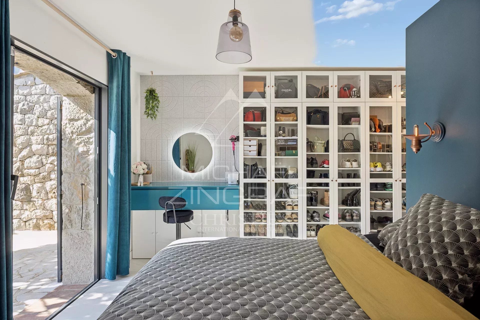 Bedroom with glass-front shoe cabinet, teal desk, round mirror and sliding door opening to a stone patio outside.