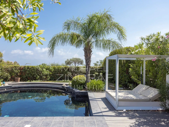 Vue d'une piscine et d'un espace détente dans un jardin luxuriant, avec des palmiers et des terrasses.