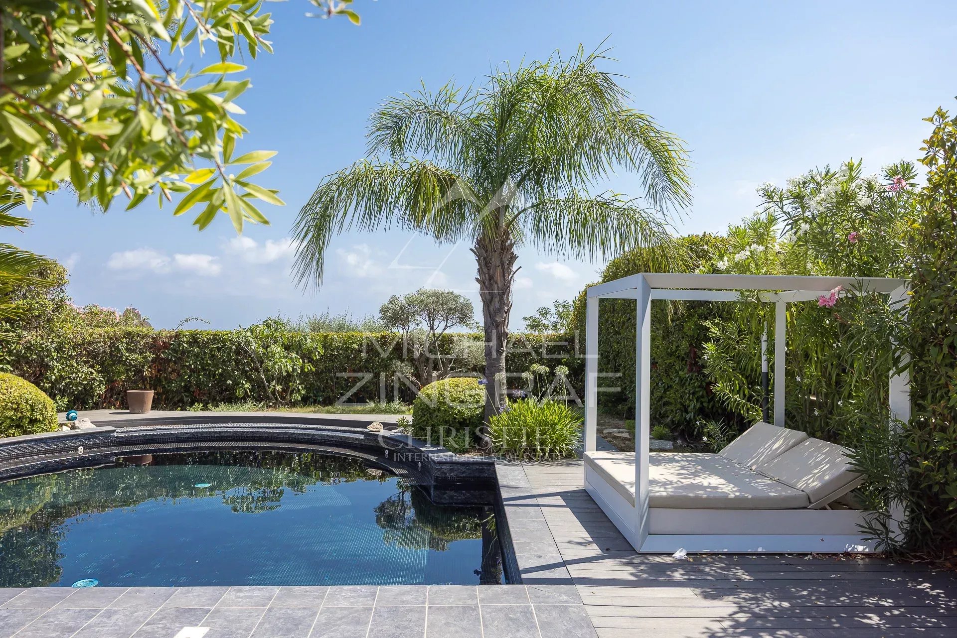 Vue d'une piscine et d'un espace détente dans un jardin luxuriant, avec des palmiers et des terrasses.