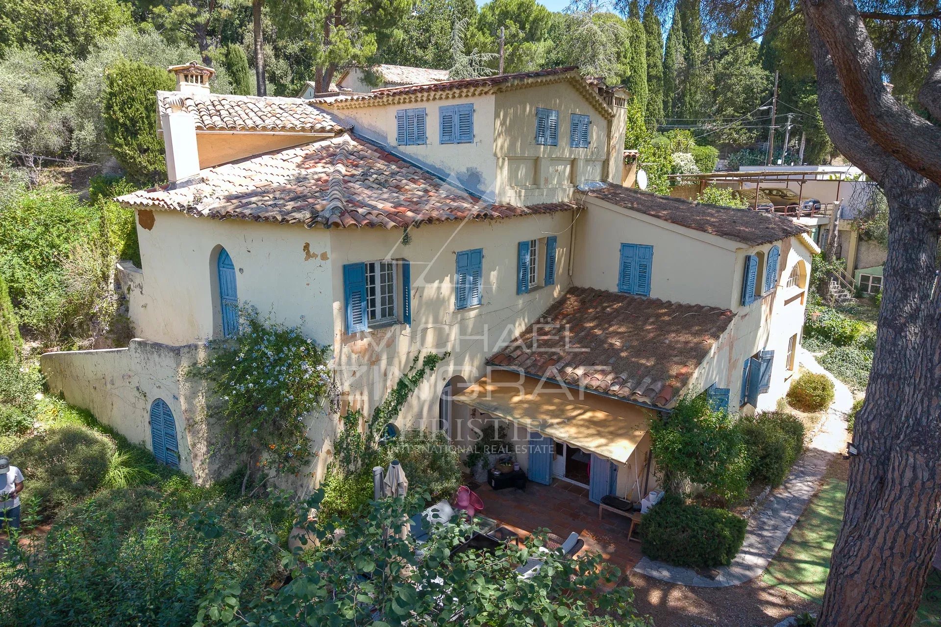 Authentique bastide du XVIIIᵉ siècle à Nice Bastide du XVIIIᵉ siècle à Nice, avec façade en pierre et volets bleus, entourée de verdure.