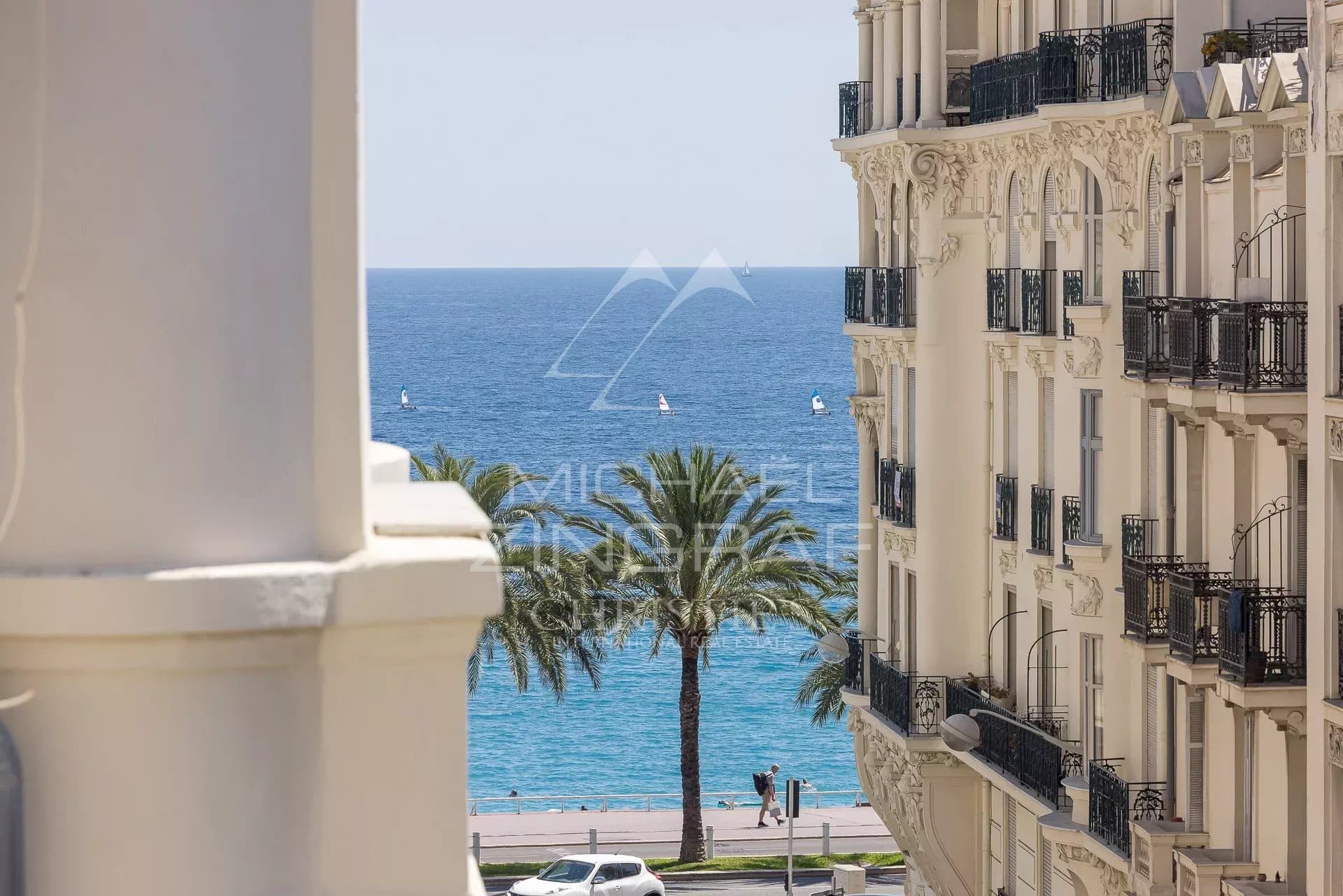 Vue latérale sur la mer Méditerranée depuis un immeuble à Nice, avec des palmiers au premier plan.