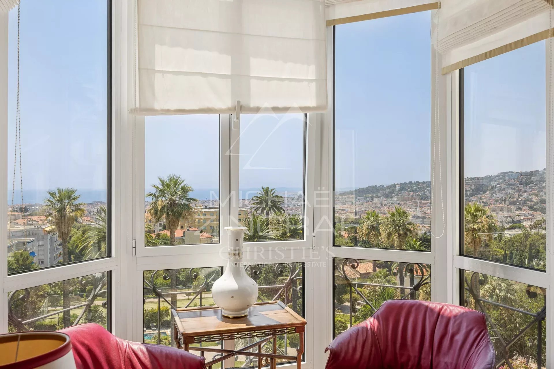 View from an apartment in Nice with red armchairs and a view of the sea and gardens.