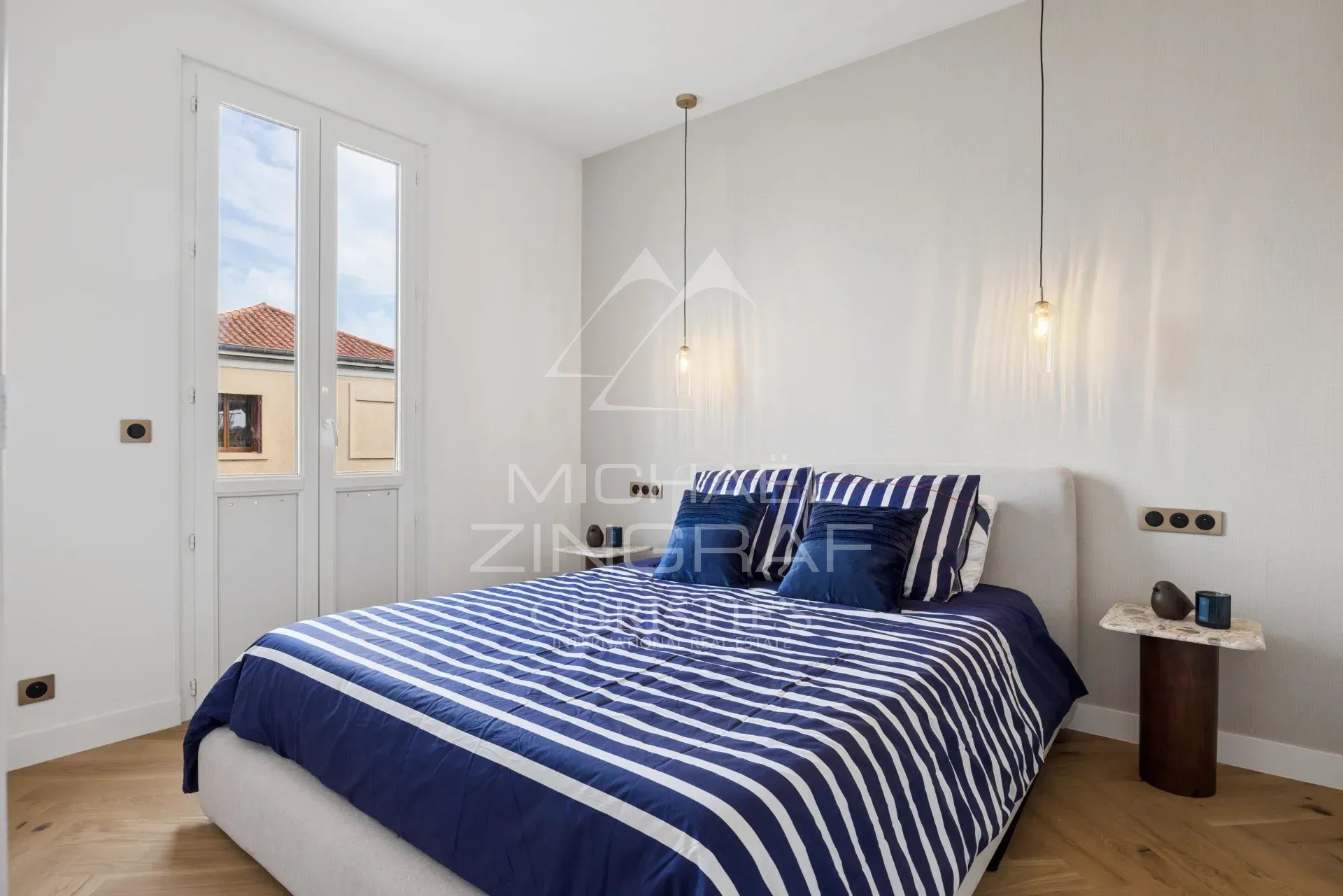 Chambre rénovée à Cros-de-Cagnes Chambre moderne avec lit à rayures bleues et blanches, fenêtres laissant entrer la lumière.