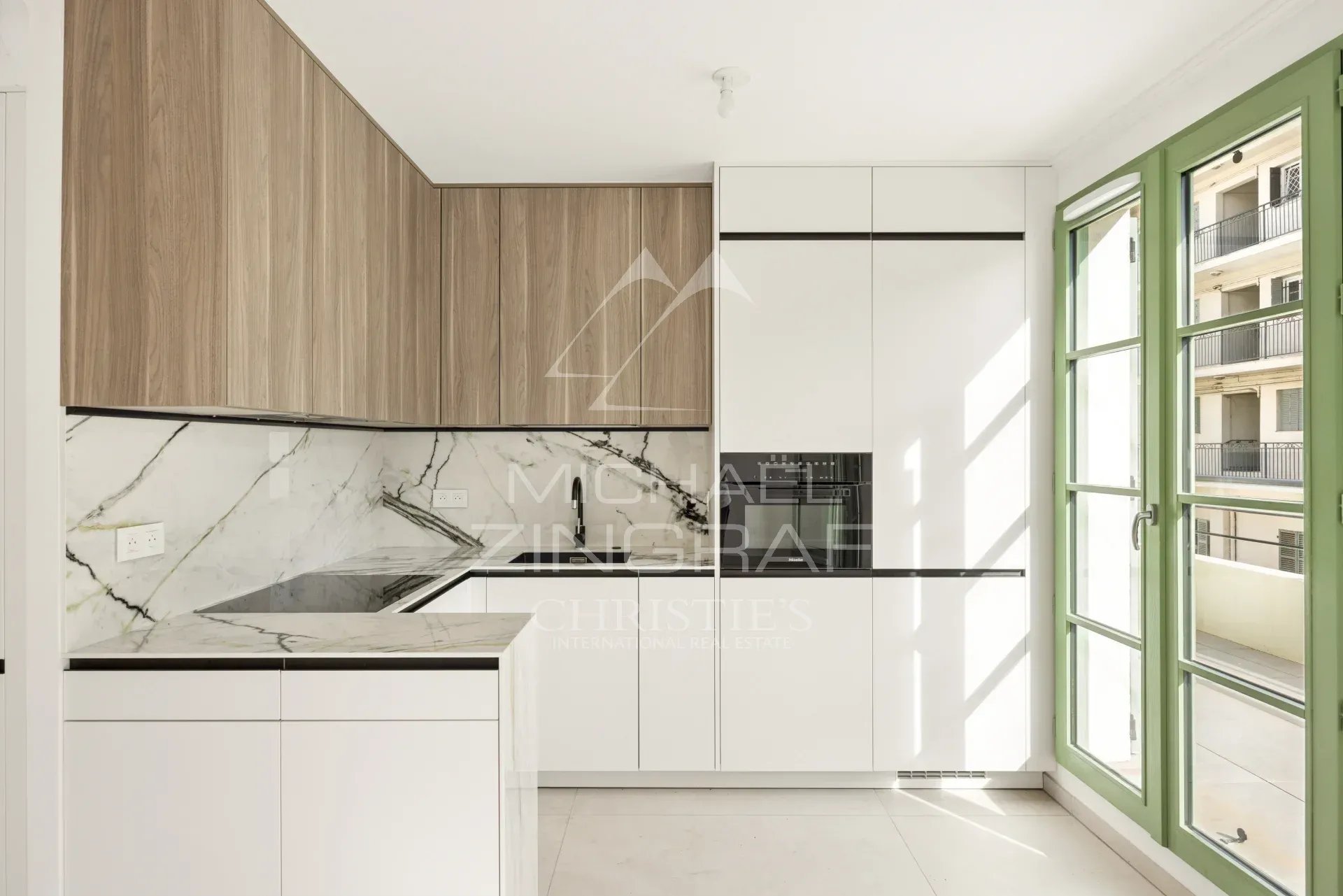 Cuisine moderne avec des meubles blancs et un plan de travail en marbre, fenêtres ouvertes sur un balcon.