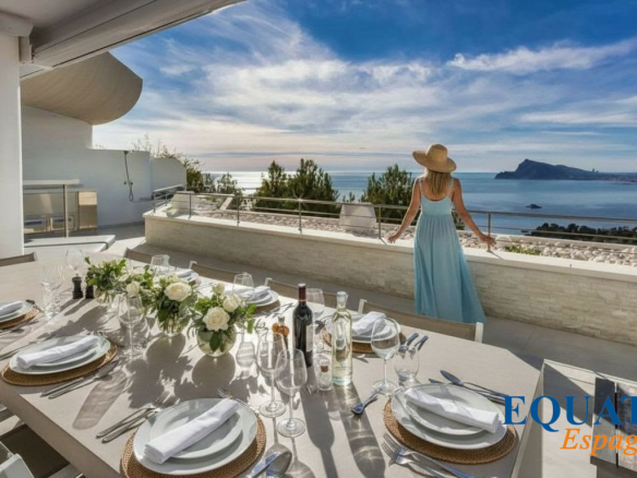 Terrace dining setup with a long table, plates, glassware, and a floral centerpiece; a woman in a blue dress and sunhat gazes at the sea.
