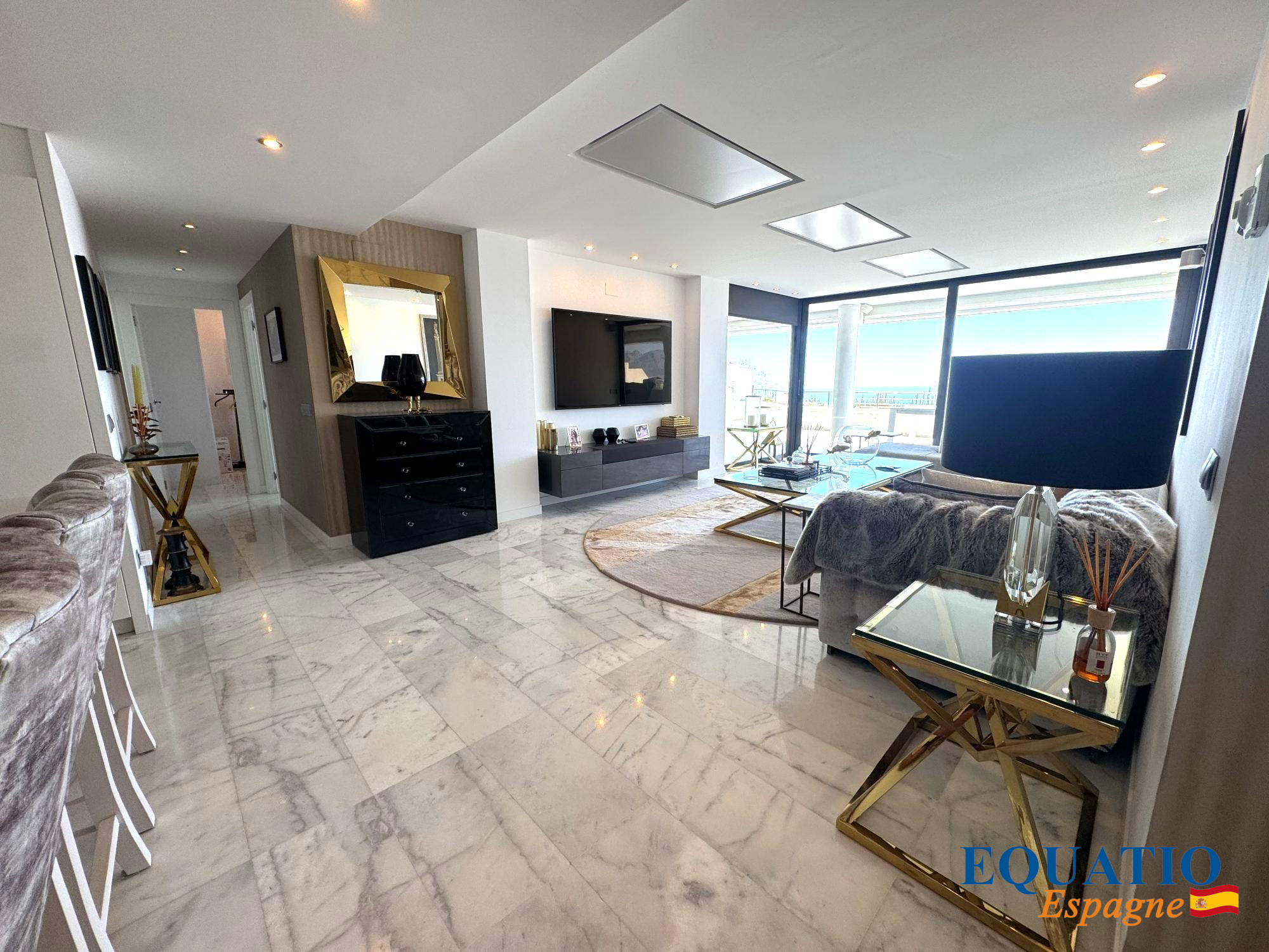 Modern living room with marble floor, wall-mounted TV, black dresser, and seating area by large windows with a balcony view.