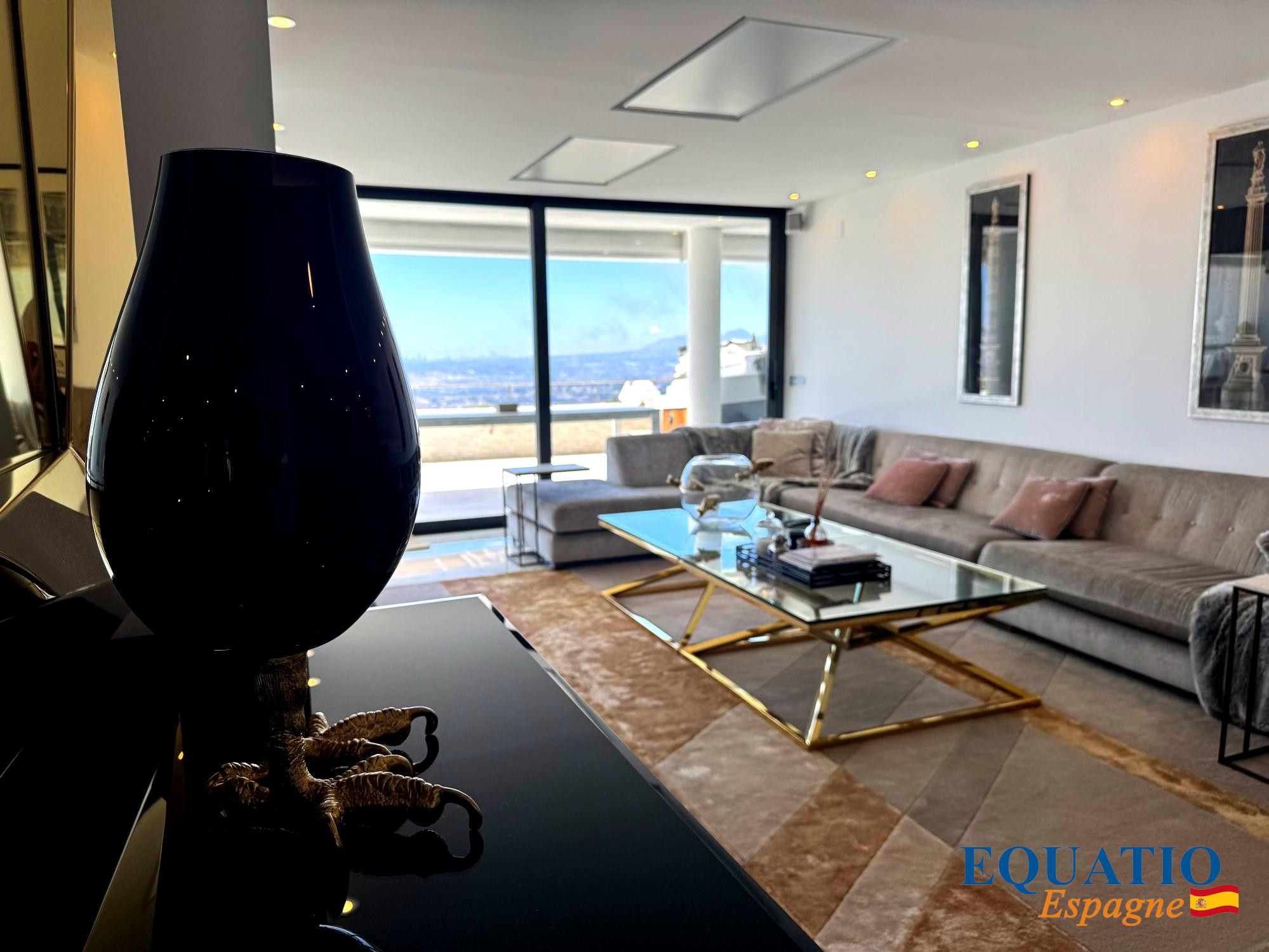 Luxurious living room with a beige sectional, glass coffee table, and a dark lamp in the foreground; large windows reveal a blue sky outside (logo 'EQUATIO Espagne' in corner).