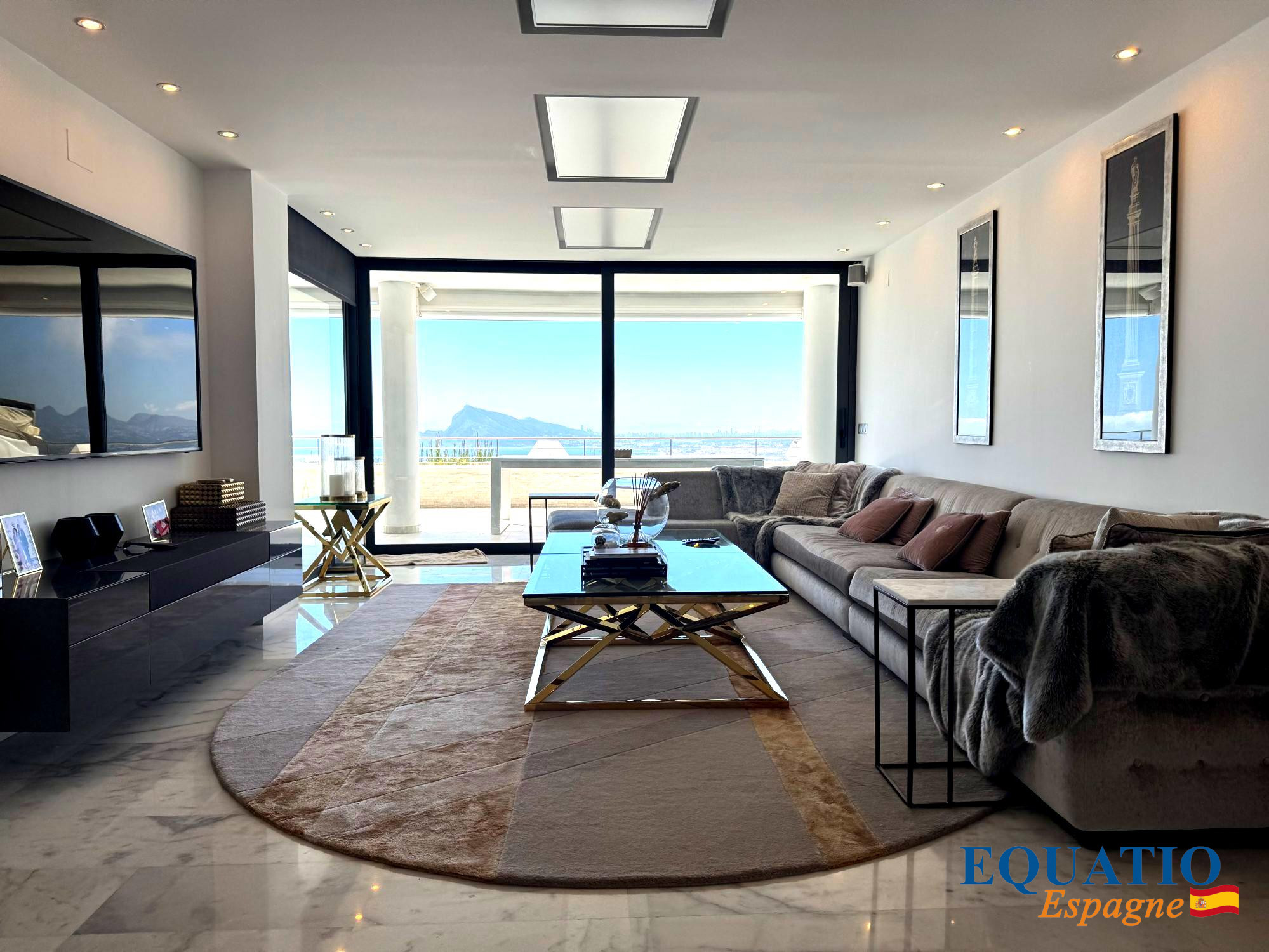 Spacious modern living room with a gray sectional, glass coffee table, and a balcony view of the sea and mountains through large windows.
