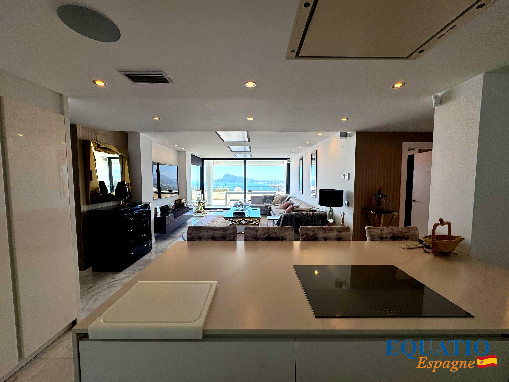 Open-plan kitchen and living area with a white island and induction cooktop in the foreground, and a bright lounge with a sea view through large windows in the distance.